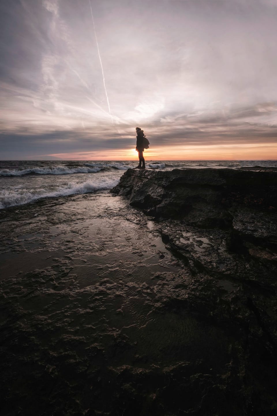 Contemplative figure on ocean rocks against sunset, embodying seizing opportunities and amplifying voice.